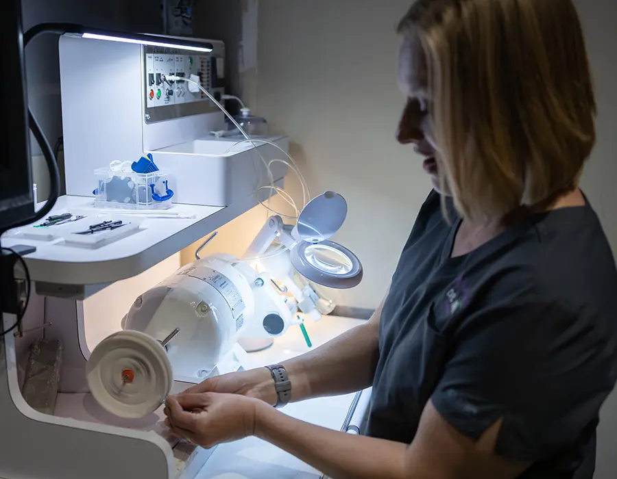 A woman in scrubs works at a laboratory station, handling equipment under a magnifying lamp with various scientific instruments and tools on the counter nearby.
