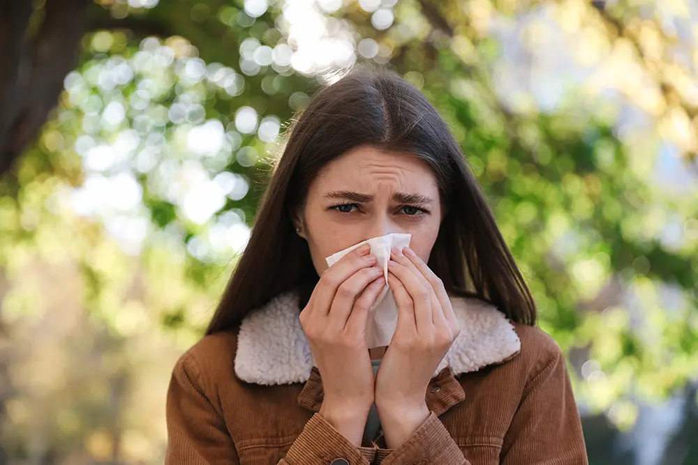 A young woman outdoors holds a tissue to her nose, appearing to sneeze or have allergies. She looks uncomfortable, surrounded by green trees and blurred sunlight in the background.