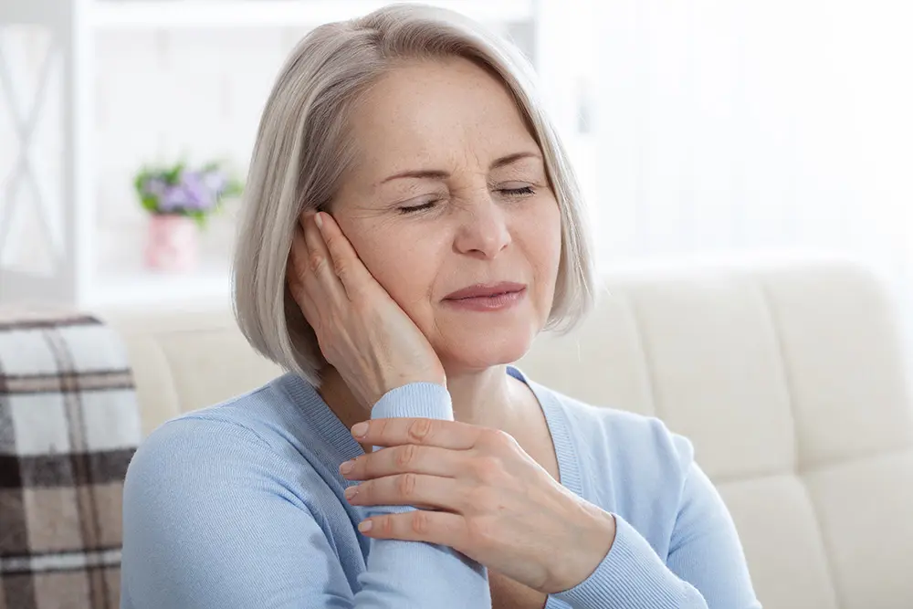 A middle-aged woman with short gray hair sits on a couch, eyes closed, and holds her hand to her ear, appearing to be in pain or discomfort. A plant and blanket are visible in the background.