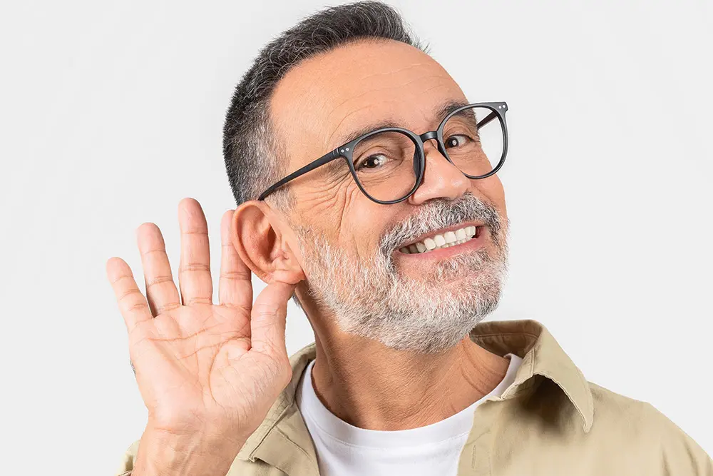 Smiling middle-aged man with glasses and a beard holding his hand to his ear as if trying to listen more closely, against a plain light background.