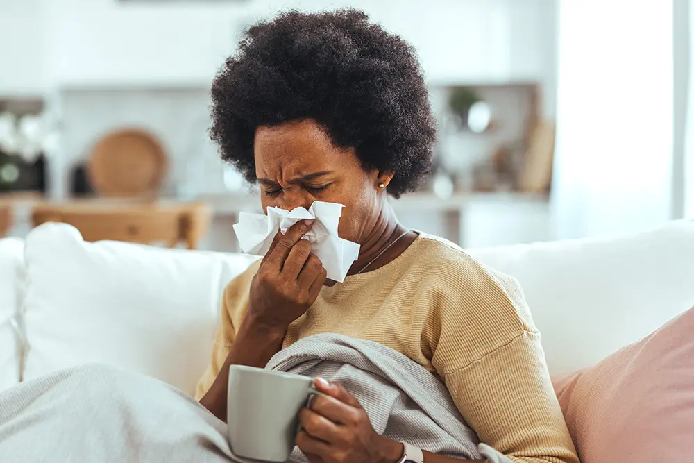 A person sits on a couch wrapped in a blanket, holding a mug and using a tissue to blow their nose. They appear unwell or sick, in a cozy home setting with a blurred kitchen in the background.