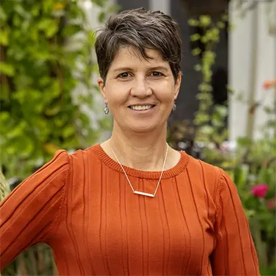 A woman with short brown hair, wearing an orange ribbed sweater and a geometric necklace, stands outdoors in front of green plants, smiling at the camera.