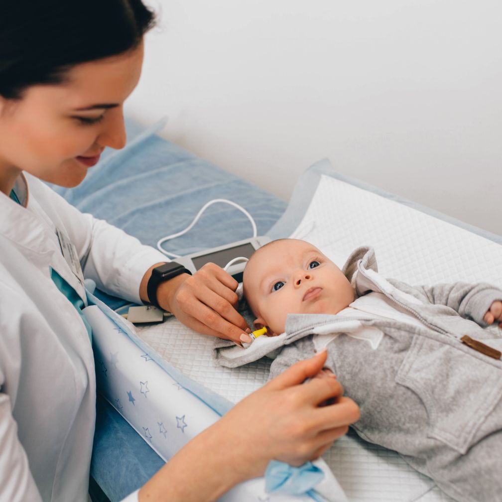 A healthcare professional tests a babys hearing using specialized equipment, gently placing sensors on the babys head as the baby lies on an exam table.
